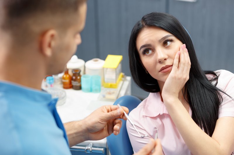 a young woman with dark hair holding her cheek as she listens to her dentist explain that she needs a root canal