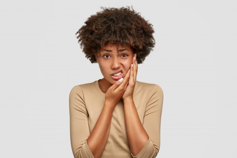 a young woman wearing a tan blouse and holding her cheek in pain because of a serious toothache